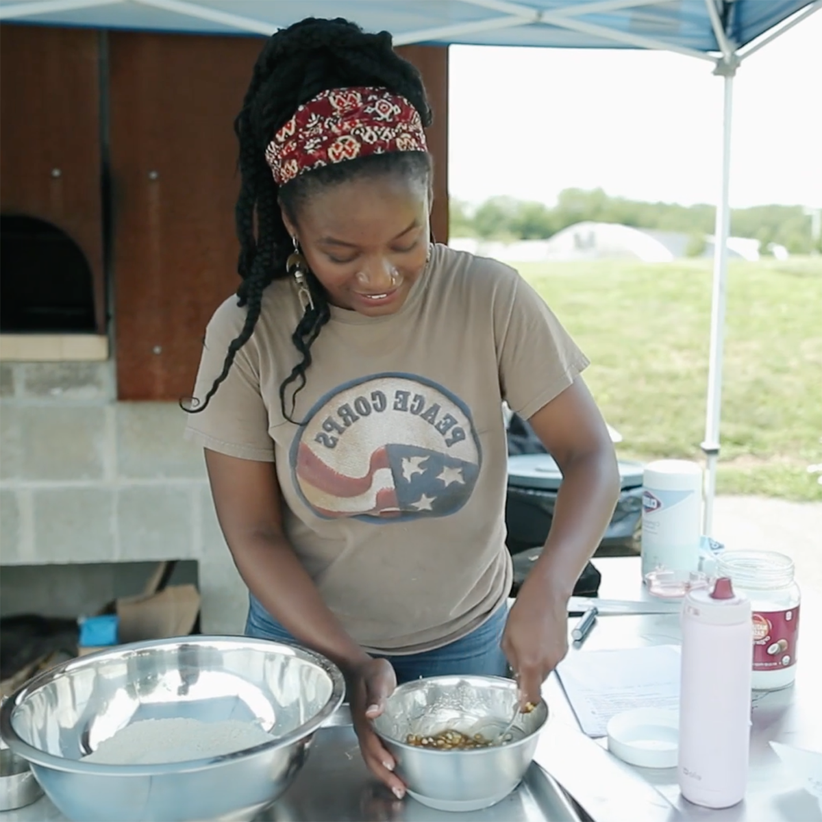 图为一位穿着和平队衬衫的年轻黑人妇女, smiling and stirring food in a bowl at the 伊甸堂校园 Bread Oven