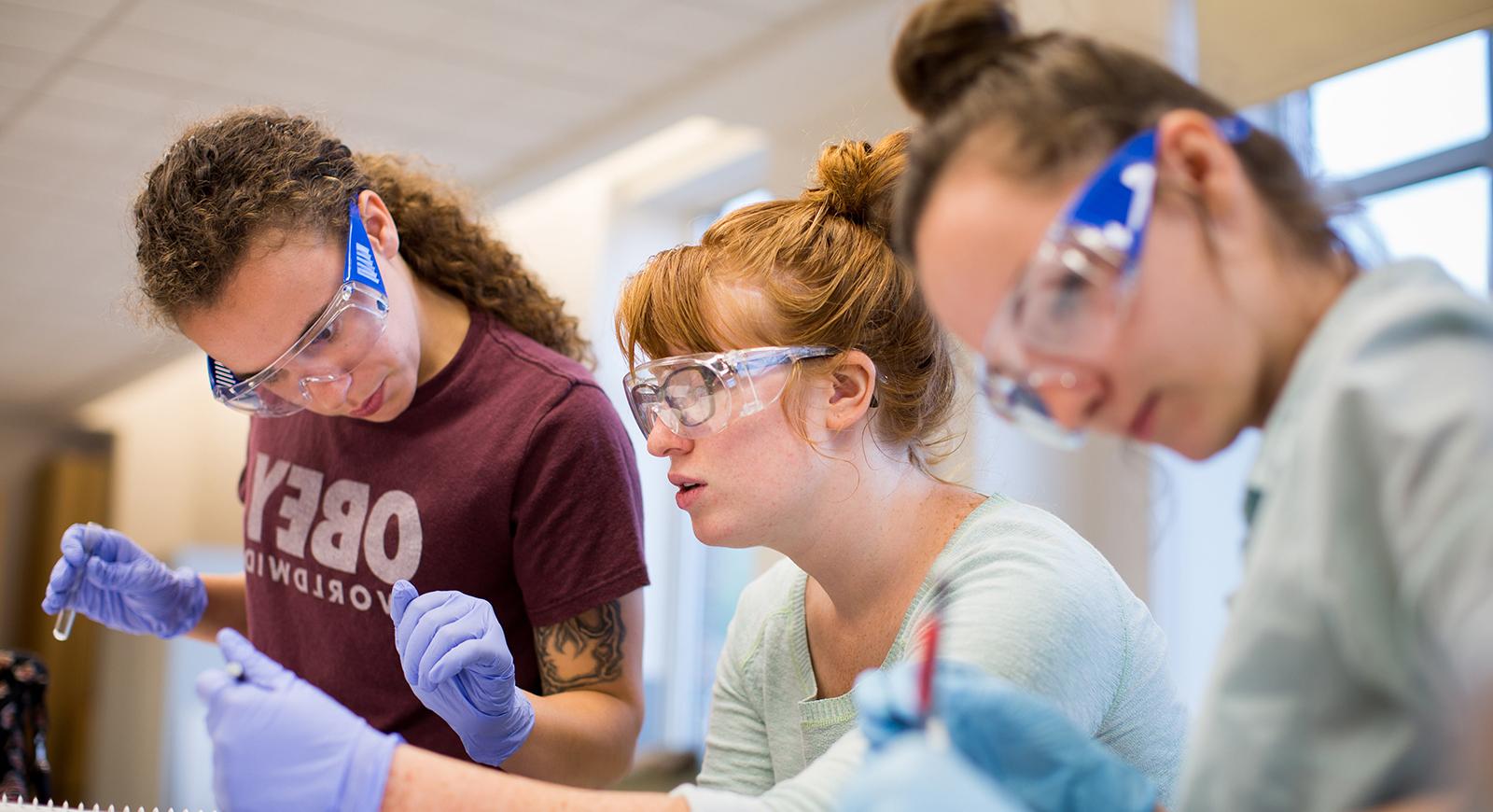 Photo of three female Chatham University biochemistry students wearing blue gloves and goggles, 拿着玻璃试管在实验室里工作. 