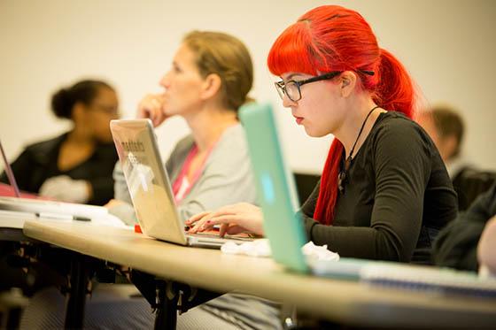 照片中的学生戴着眼镜，头发鲜红, working on her laptop in a Chatham University lecture hall