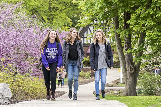 Photo of three female Chatham University walking to class on 足球波胆平台 during the spring, with green trees on colorful blossoms. 